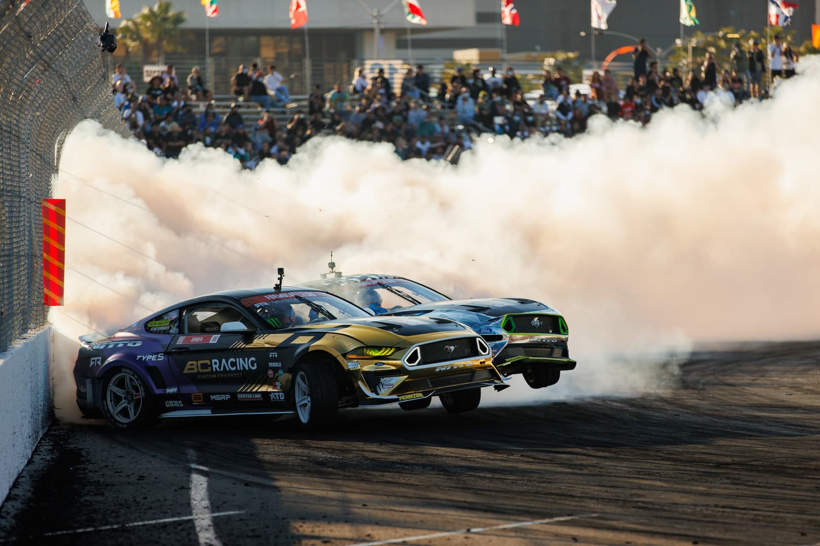 Two drift Mustangs battling through smoke in front of a crowd