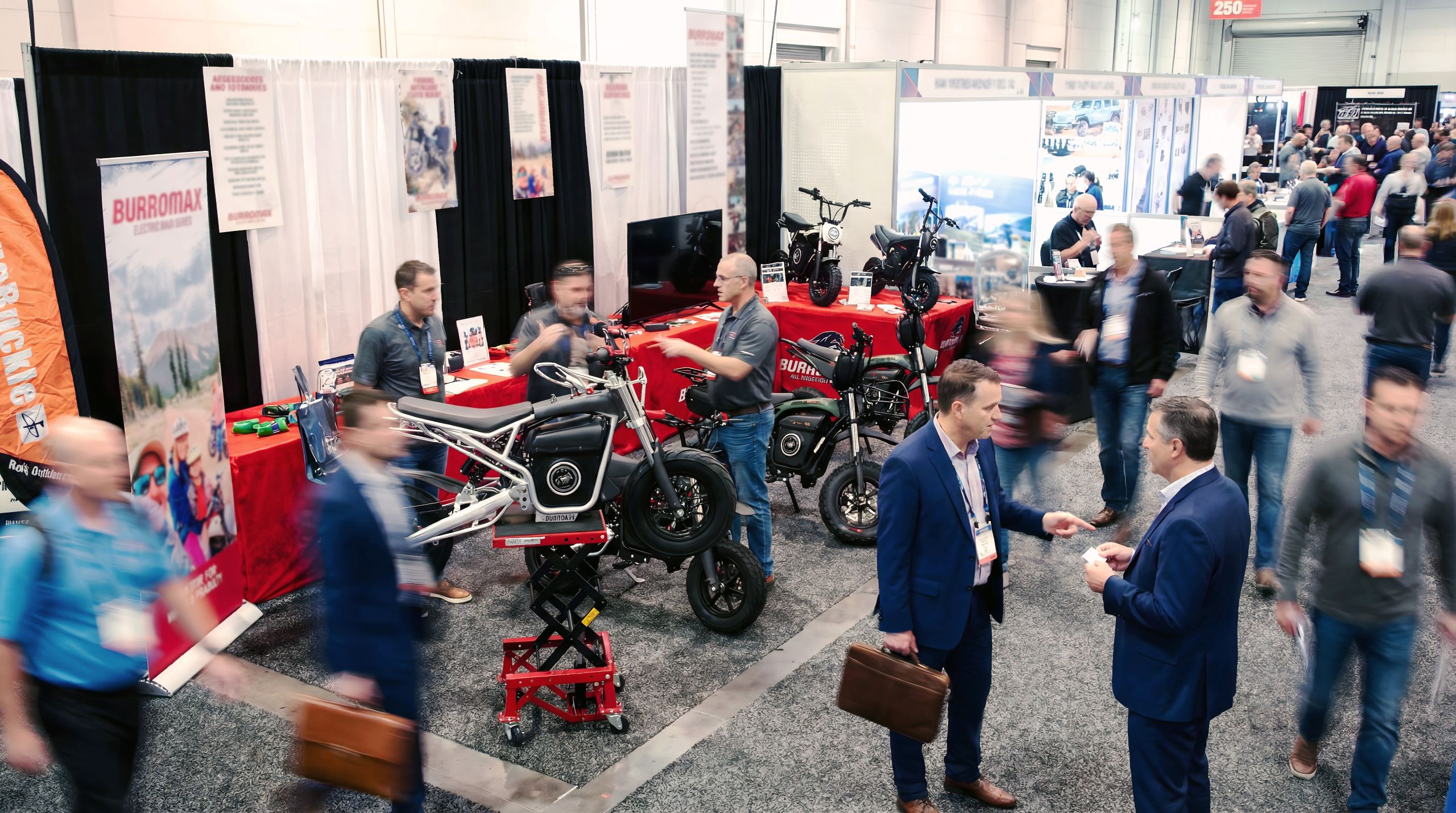 Wide shot of the Burromax booth at a trade show featuring various electric mini bikes and attendees walking by.