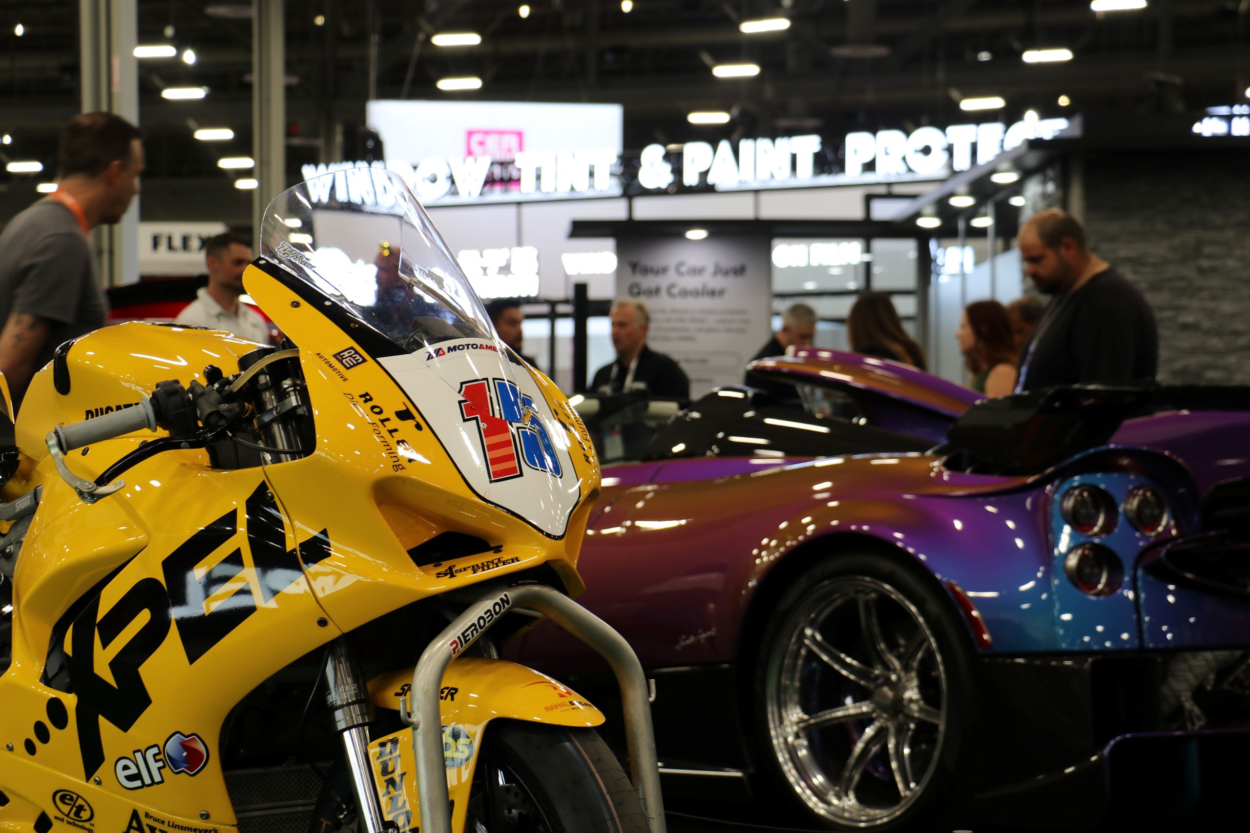 A yellow Rahal Ducati MotoAmerica racing motorcycle sponsored by Roller Die + Forming on display next to a purple hypercar