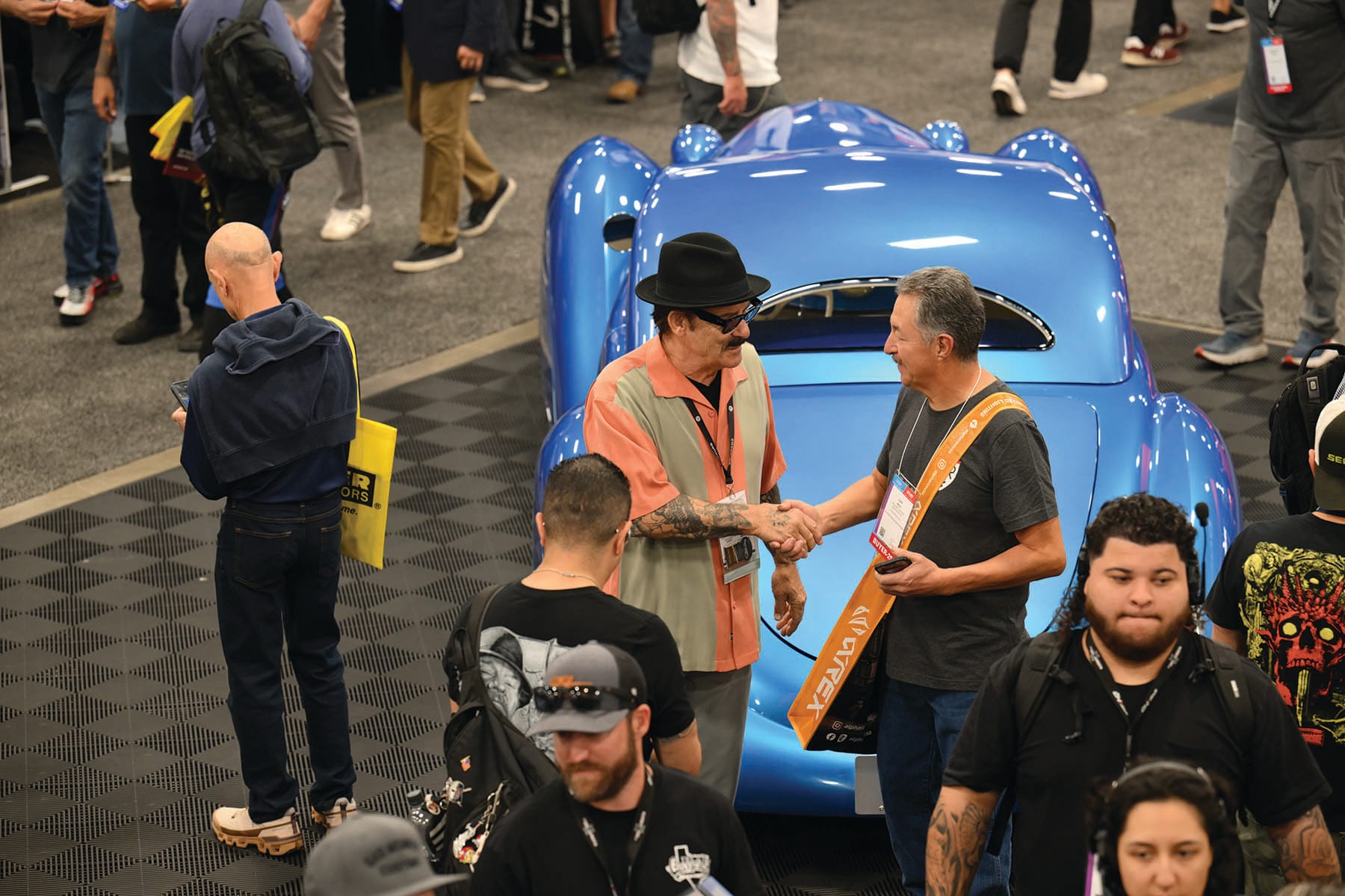 Two men shaking hands in front of a glossy blue custom car in a busy show floor aisle
