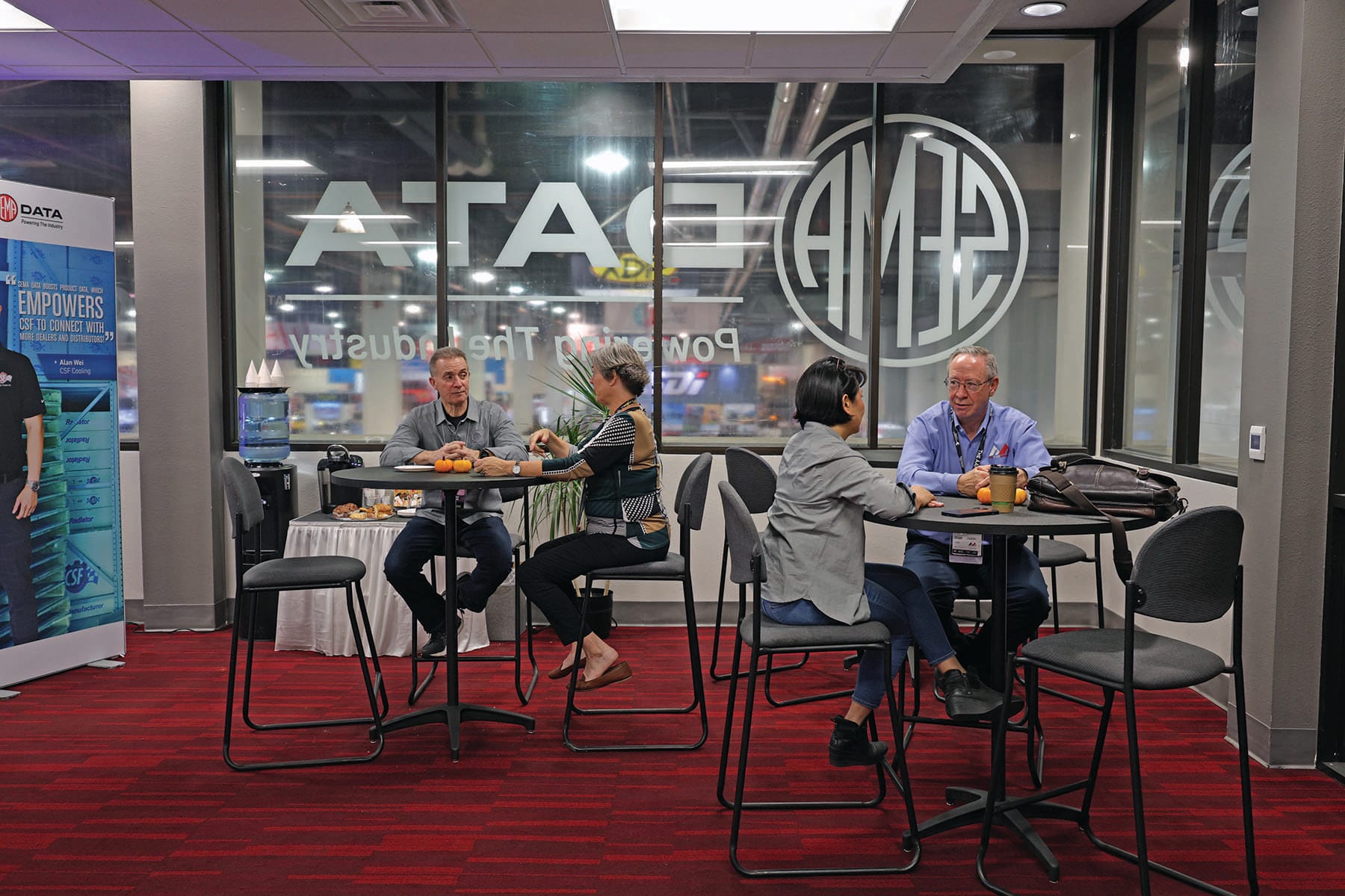 Four people talking at two small tables in the SEMA Data lounge, with the SEMA Data logo on the windows behind them