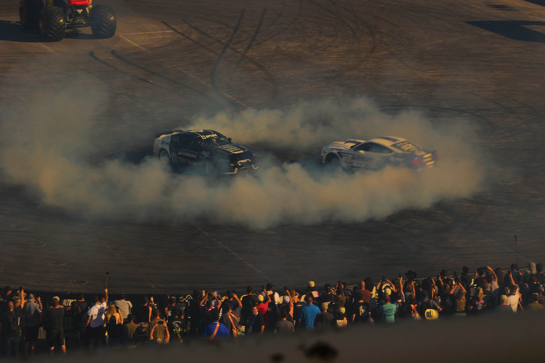 Two cars drifting and causing smoke on a closed course with crowd looking